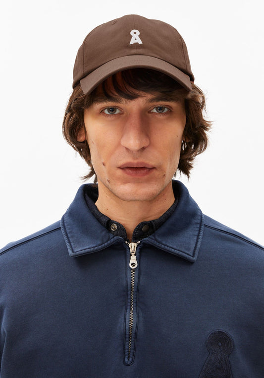 Man wearing a brown cap with a logo and a navy blue jacket on a white background