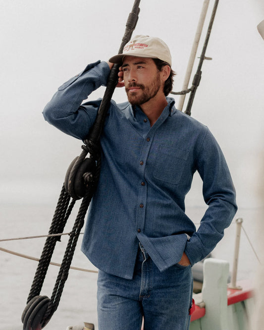CROPPED PHOTO OF MALE MODEL, LEANING ON SAILBOAT LINE, WEARING DARK BLUE DENIM SHIRT AND JEANS, WHITE CAP, SMILING, DARK SHORT HAIR AND FACIAL HAIR