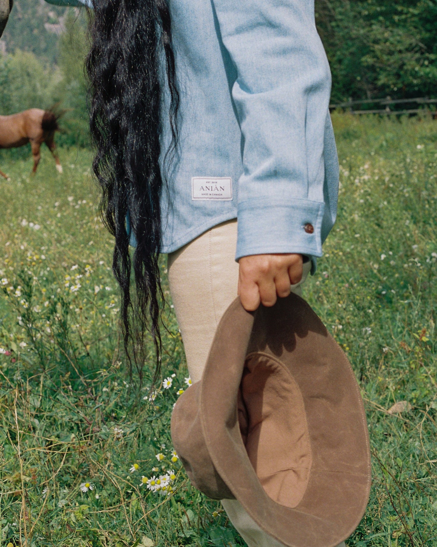 MODEL WEARS LIGHT BLUE DENIM SHIRT IN FIELD, HOLDING HAT WITH LONG BLACK HAIR - HORSE IN BACKGROUND