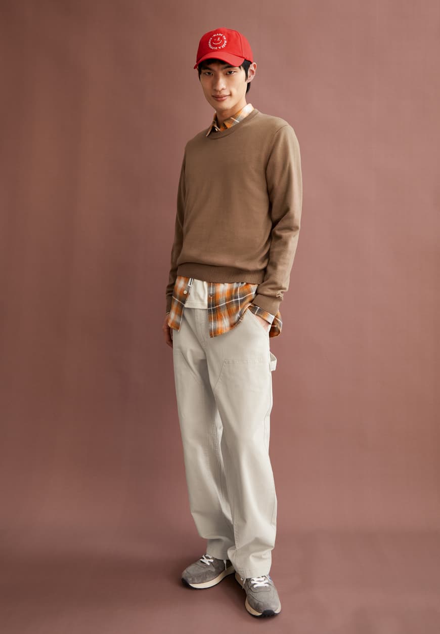 Men's casual outfit by ARMEDANGELS with brown sweater, plaid shirt, beige pants, red cap, and gray sneakers