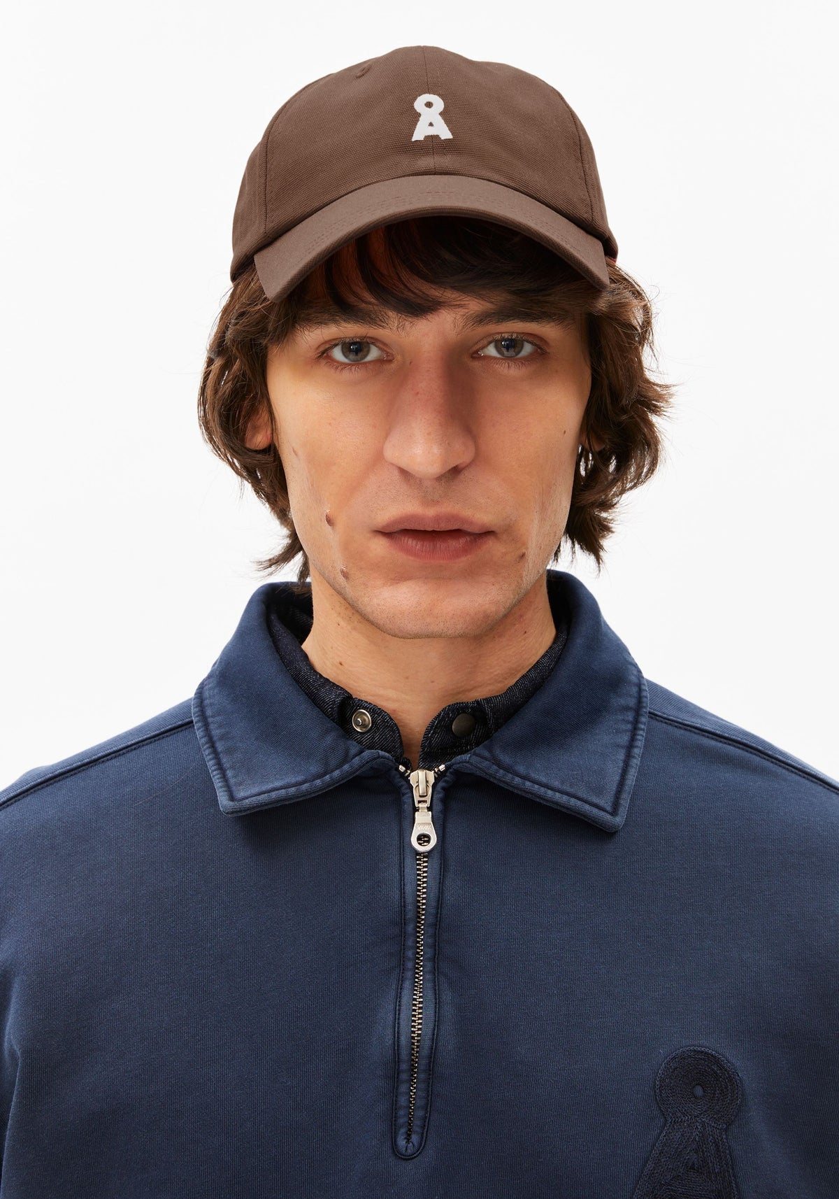 Man wearing a brown cap with a logo and a navy blue jacket on a white background