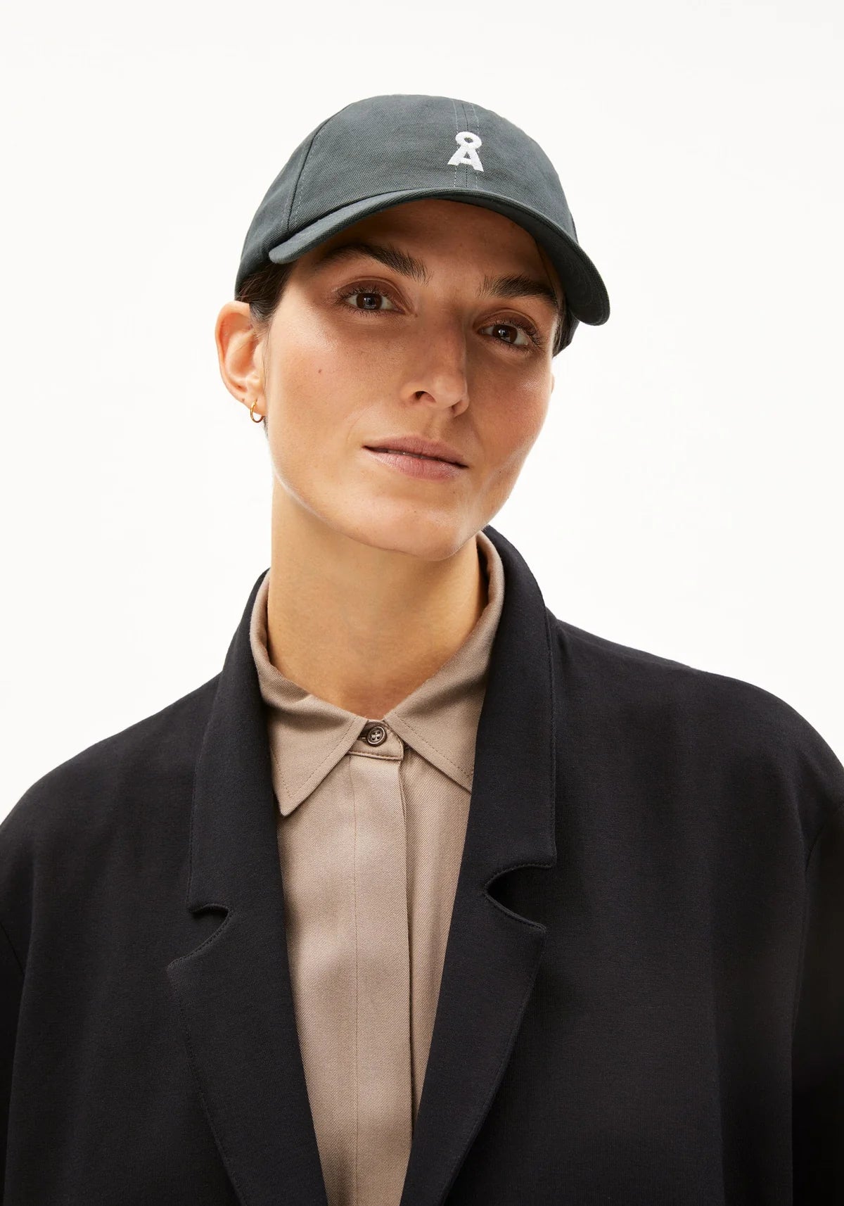 Person wearing a dark cap with a logo, black coat, and beige shirt on a white background
