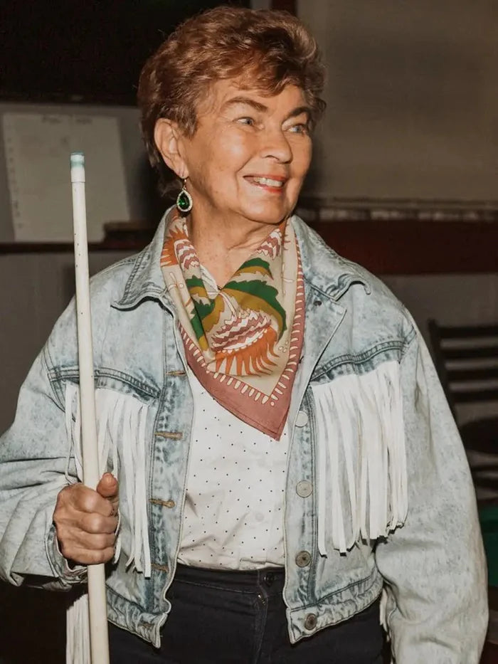 Woman in a denim jacket with fringe holding a pool cue, wearing a patterned scarf.