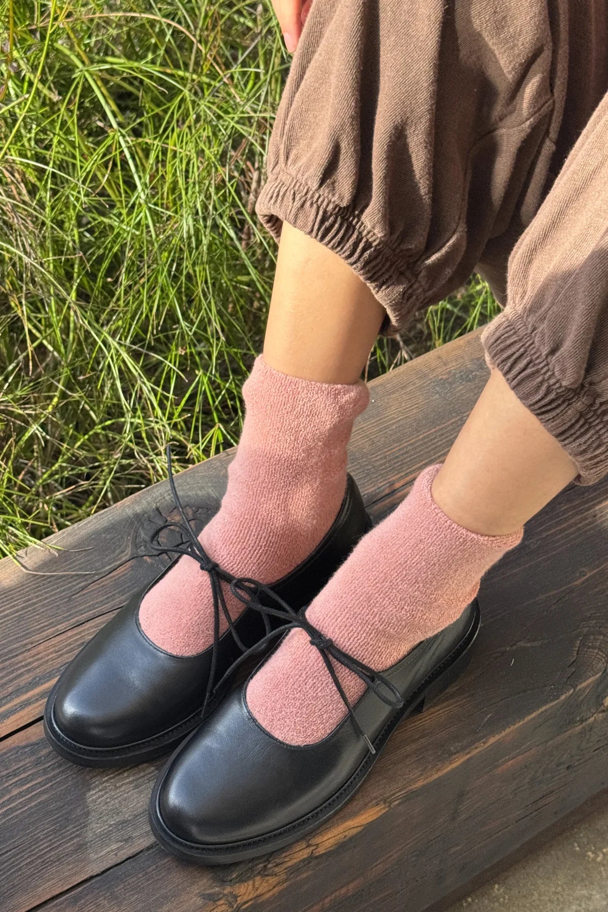 Person wearing black shoes and pink socks sitting on a wooden bench with grass in the background
