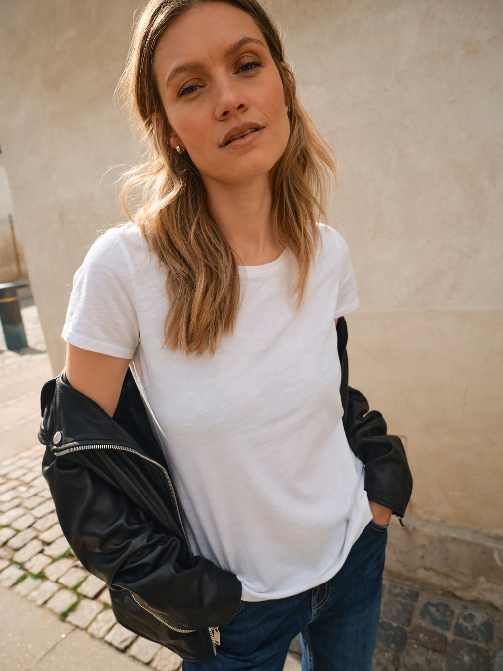 Woman wearing a white t-shirt and black leather jacket against a beige wall.