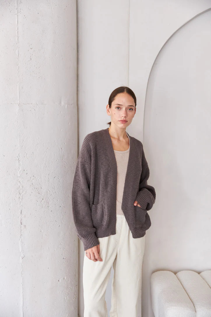Woman wearing a dark gray cardigan over a white top and pants, standing against a white wall.
