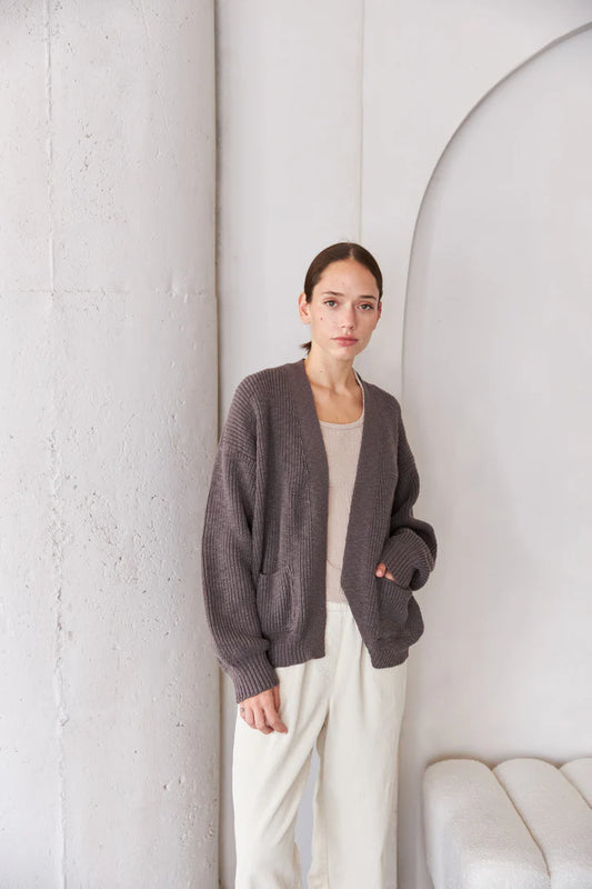 Woman wearing a dark gray cardigan over a white top and pants, standing against a white wall.