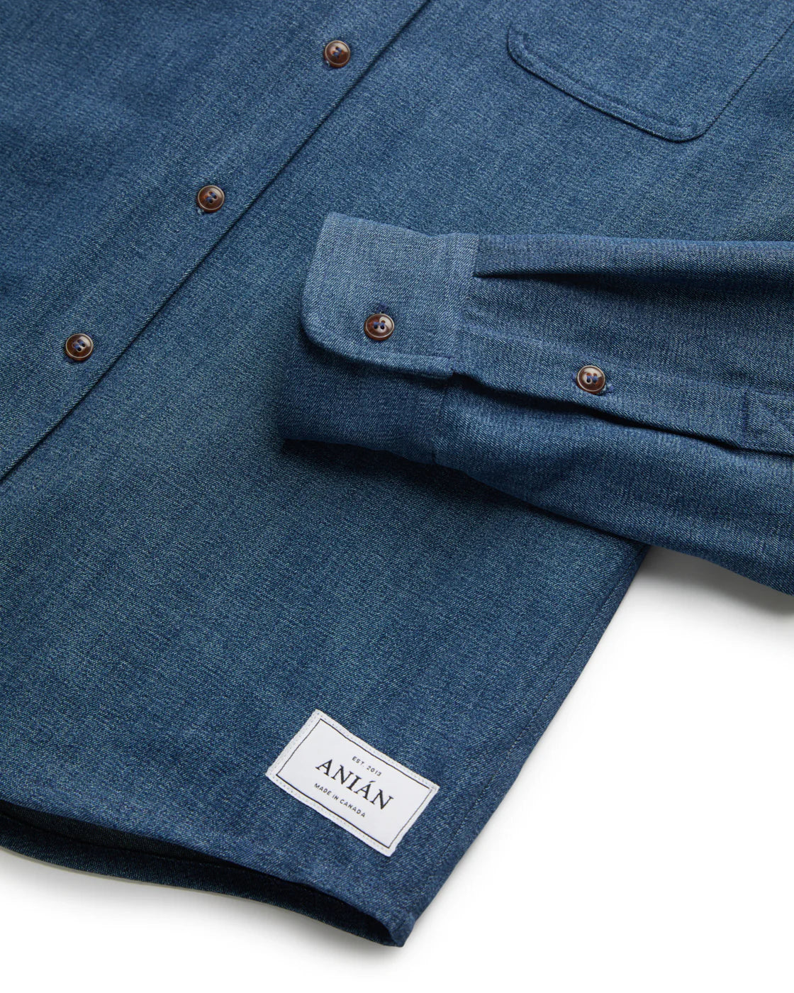 CLOSE UP OF ONE CUFF HEM OF DARK BLUE DENIM SHIRT LAID FLAT AGAINST WHITE BACKGROUND 