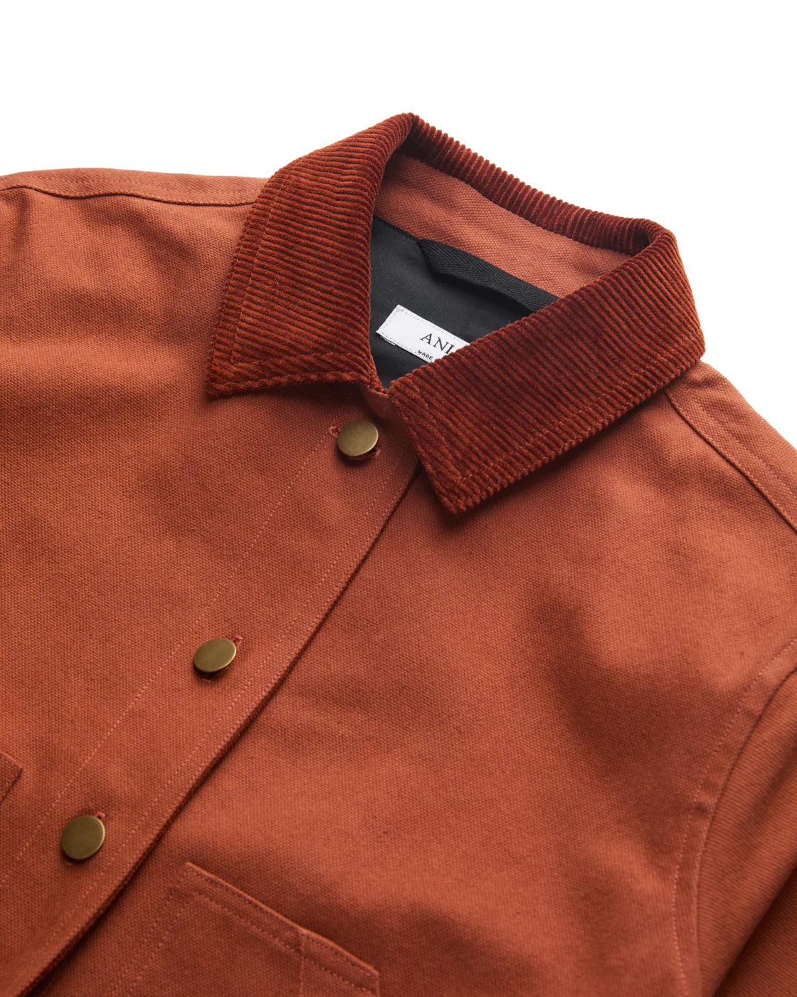 RED-ORANGE BARN COAT CLOSE UP FLAY LAY OF CORDUROY COLLAR, CANVAS BODY, BRASS BUTTONS, AGAINST A WHITE BACKGROUND