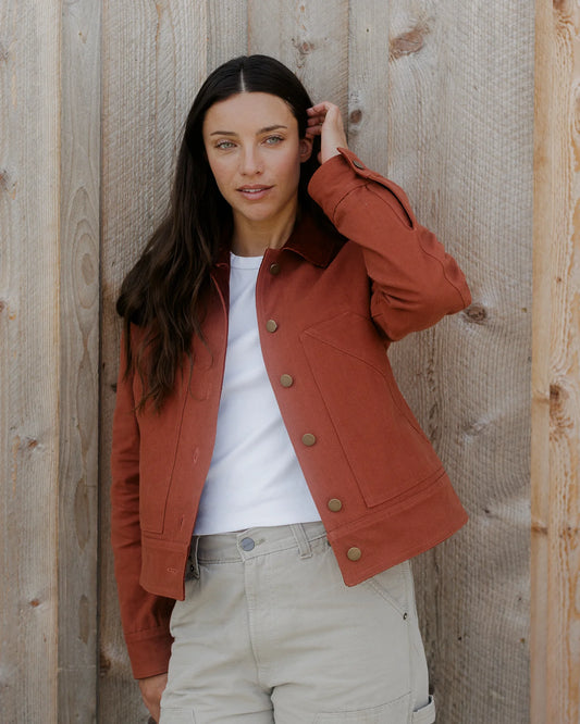 3/4 MODEL SHOT IN RED-ORANGE BARN COAT, WHITE T-SHIRT, AND TAN KHAKI PANTS, LEANING AGAINST WOOD WALL BACKGROUND, LEFT HAND IN HAIR