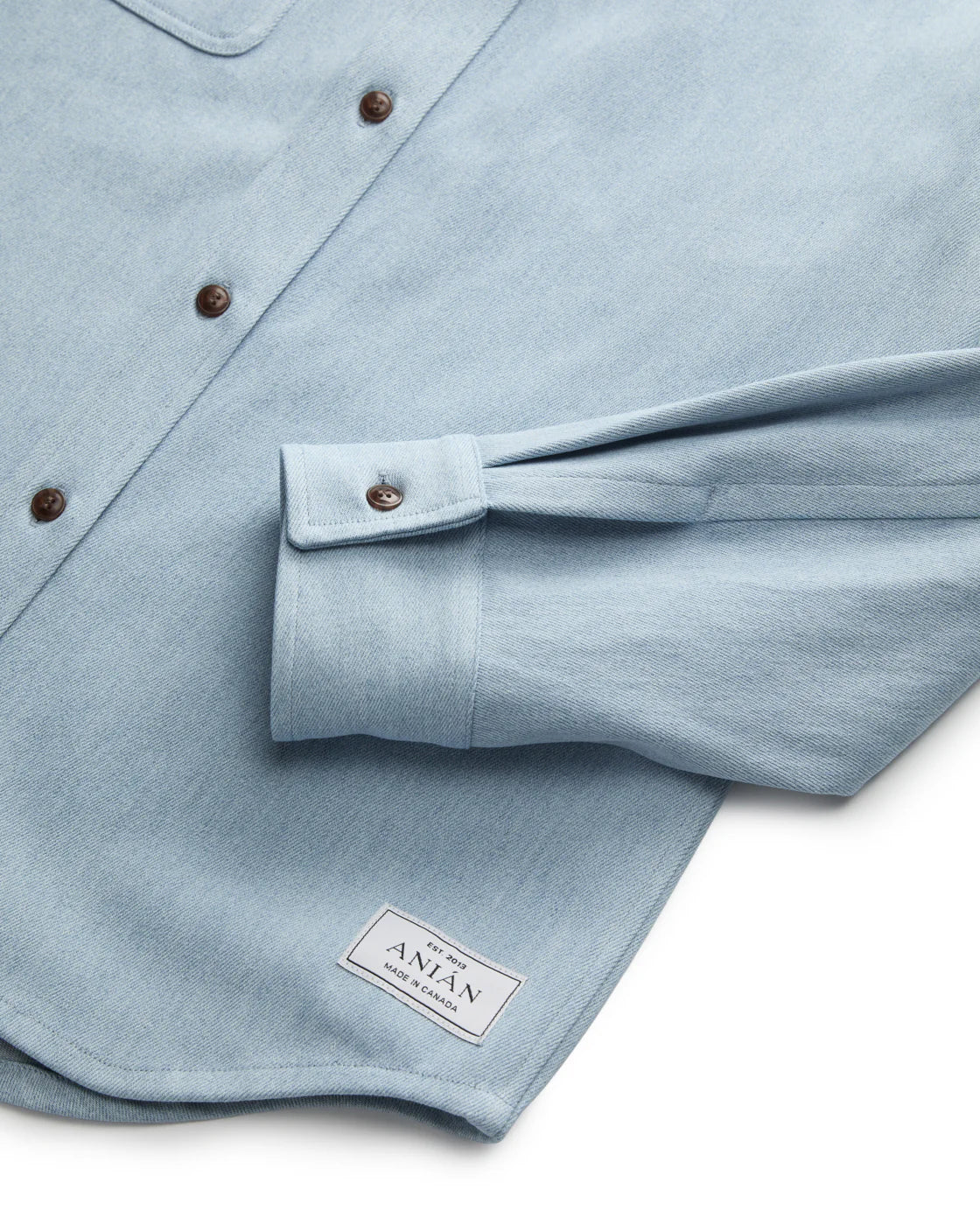 CLOSE UP OF ONE CUFF HEM OF LIGHT BLUE DENIM SHIRT LAID FLAT AGAINST WHITE BACKGROUND 