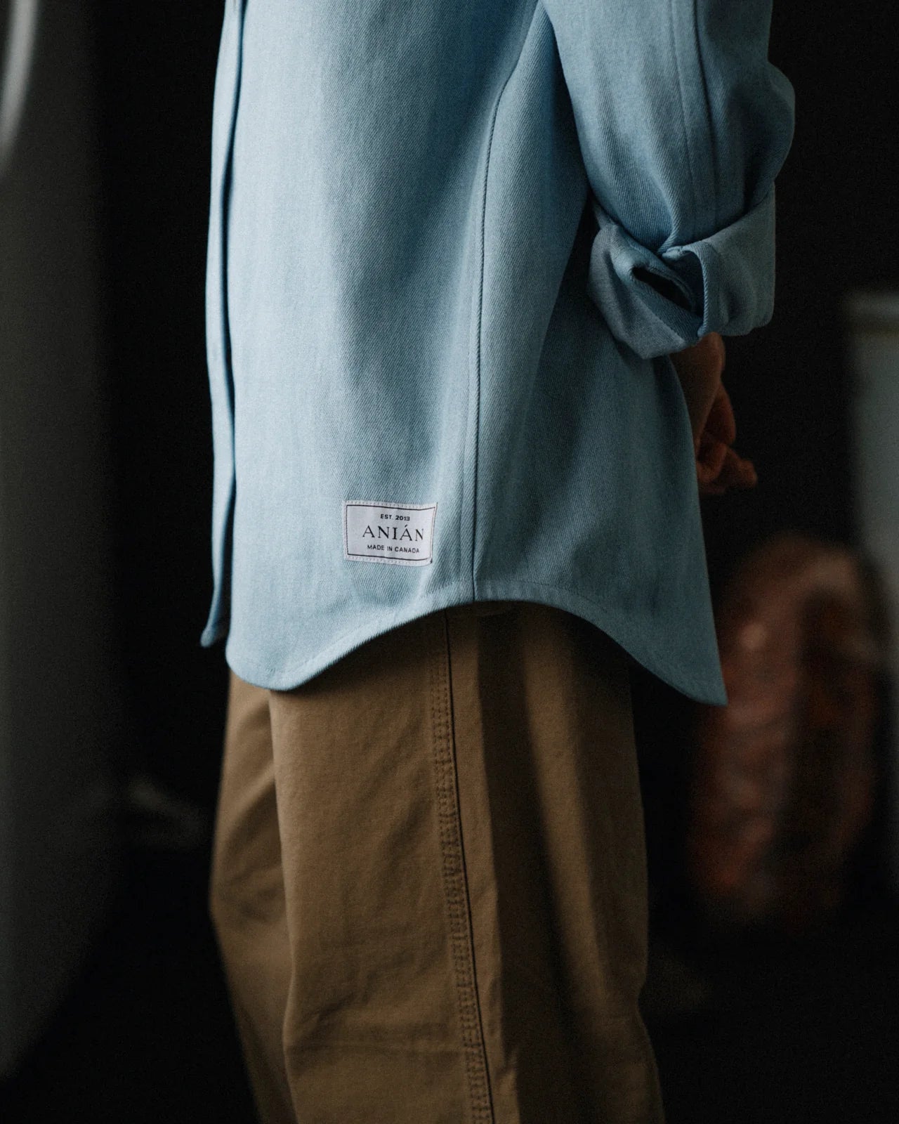 MID-SHOT OF MODEL WEARING A LIGHT BLUE DENIM SHIRT WITH CUFFED SLEEVES AGAINST A DARK BACKGROUND