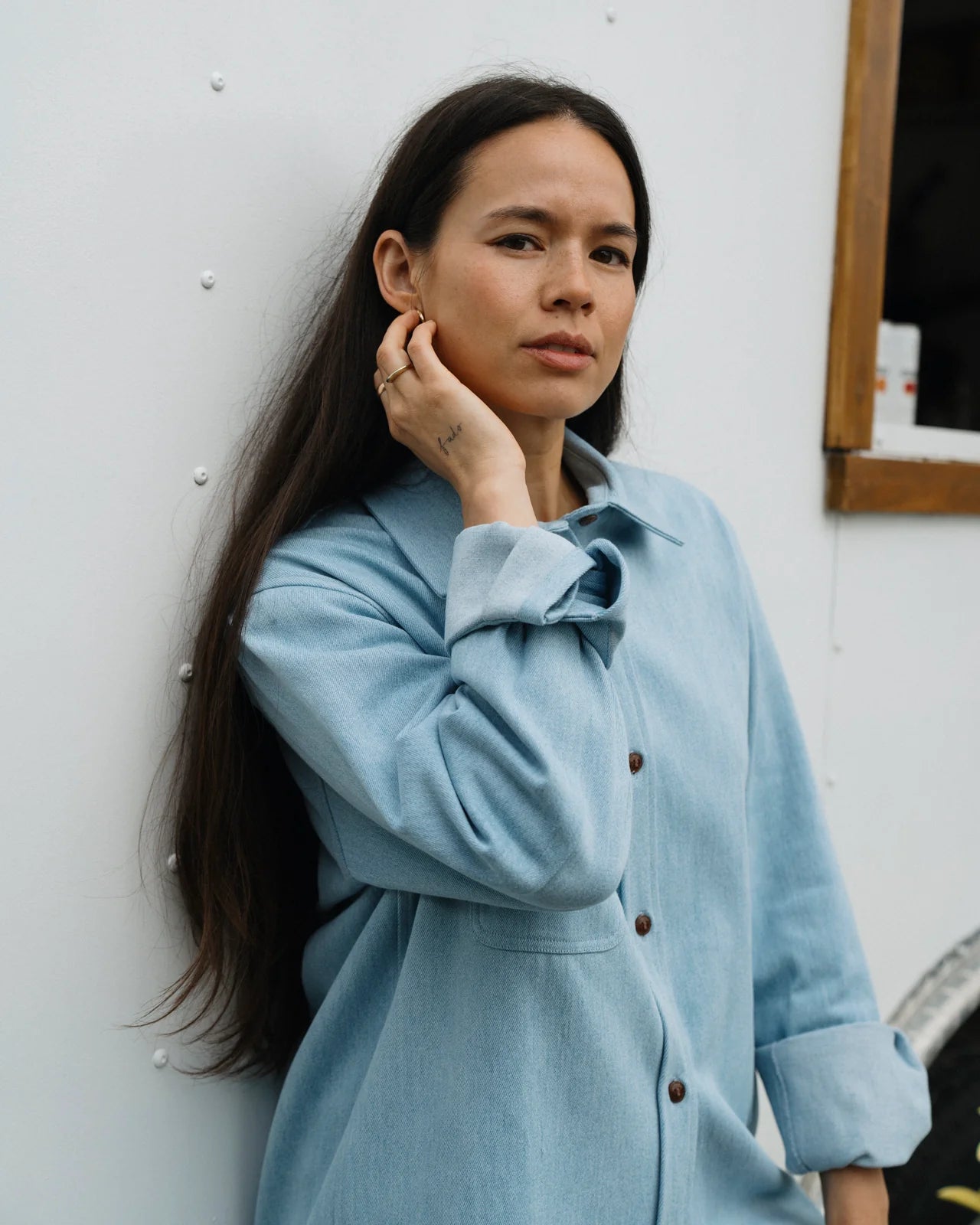 FEMALE MODEL, CLOSEUP SHOT, WEARING A LIGHT BLUE DENIM SHIRT WITH CUFFED SLEEVES AND LEANING AGAINST WHITE TRAILER