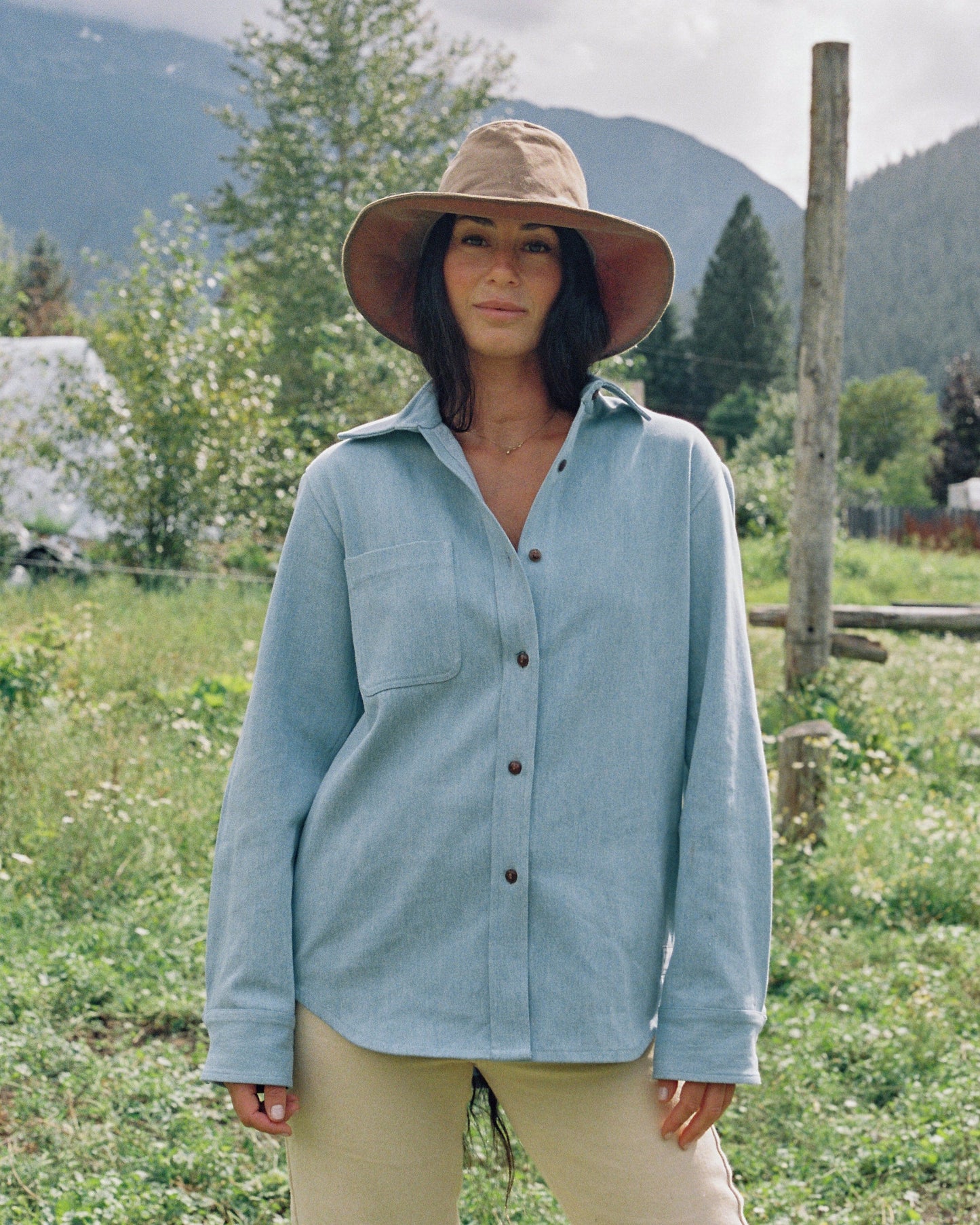 MODEL WEARS LIGHT BLUE DENIM SHIRT IN MEADOW WITH AN UNBUTTONED COLLAR AND WEARING A BIG HAT.