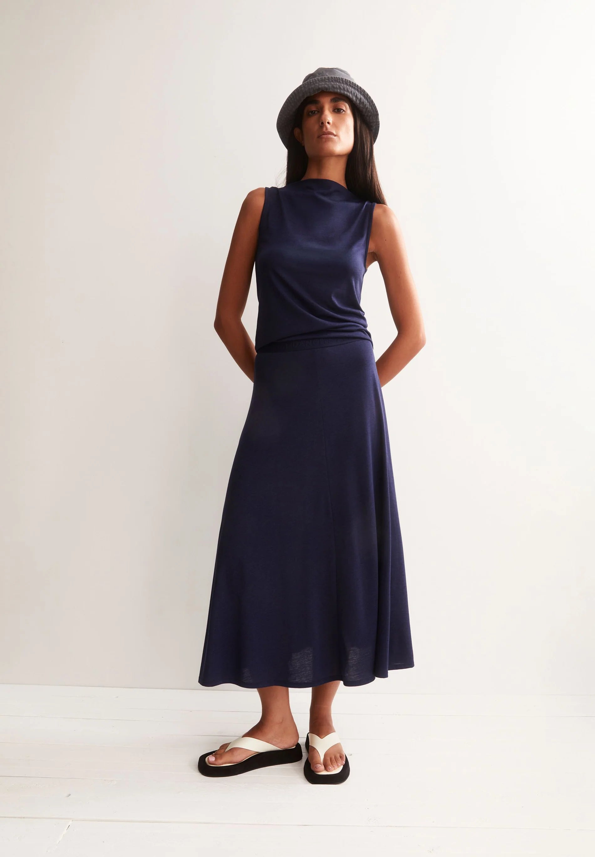 MODEL WEARING NAVY TOP AND SKIRT, GREY BUCKET HAT AND WHITE SANDALS  AGAINST A WHITE BACKGROUND