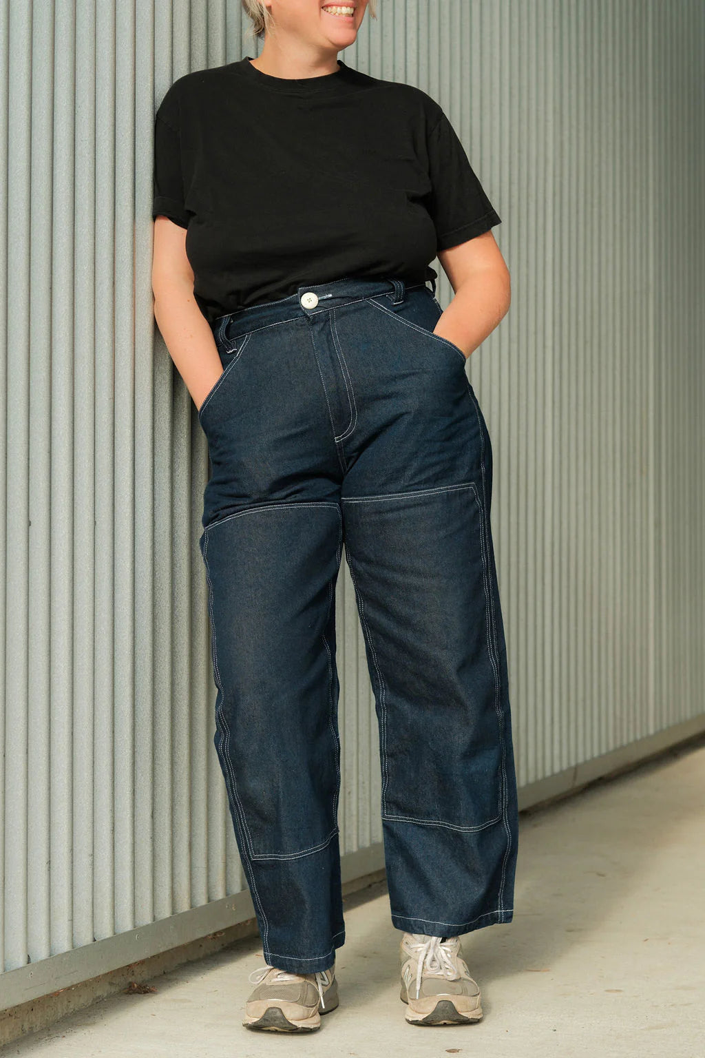 Person wearing a black t-shirt and blue jeans standing against a corrugated metal wall.