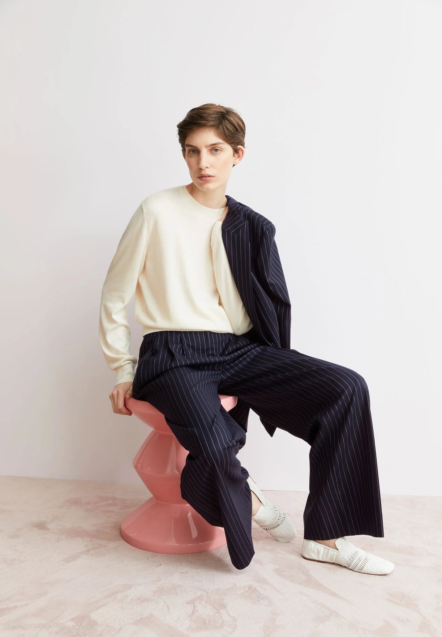 Lifestyle shot of woman sitting wearing ARMEDANGELS Canada FINE Oatmilk long sleeve sweater and a striped suit