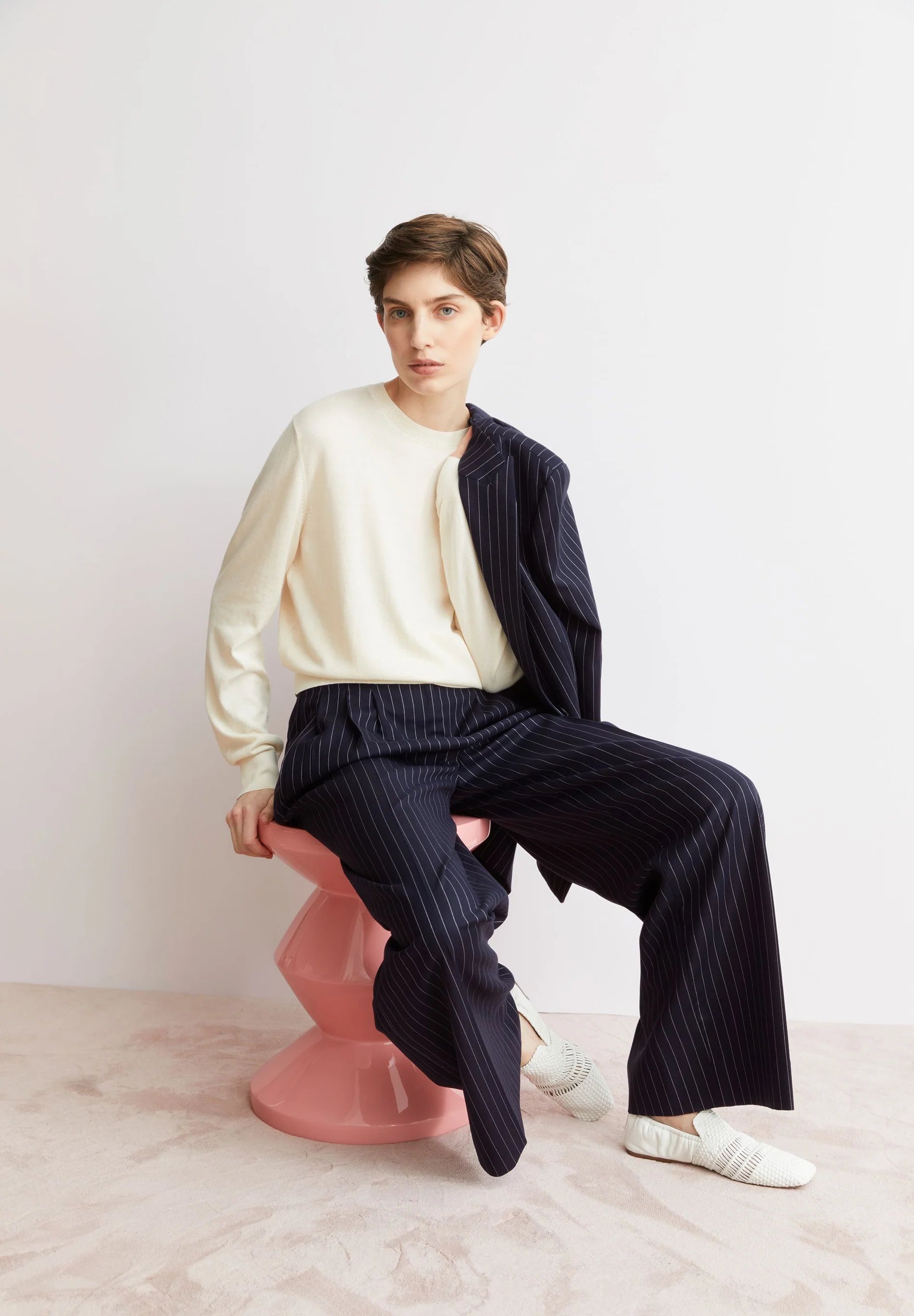 Lifestyle shot of woman sitting wearing ARMEDANGELS Canada FINE Oatmilk long sleeve sweater and a striped suit